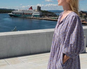 Puede incluir: Un vestido holgado de largo hasta la pantorrilla en tela de color morado claro con un estampado floral pequeño. La prenda cuenta con mangas abullonadas voluminosas, cintura fruncida, escote redondo sencillo con lazo y bolsillos laterales.