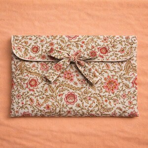 Op de afbeelding: Een clutch met bloemenpatroon en striksluiting. De clutch heeft een cr&egrave;mekleurige achtergrond met rode en roze bloemen en groene bladeren. De achtergrond is perzikkleurig.