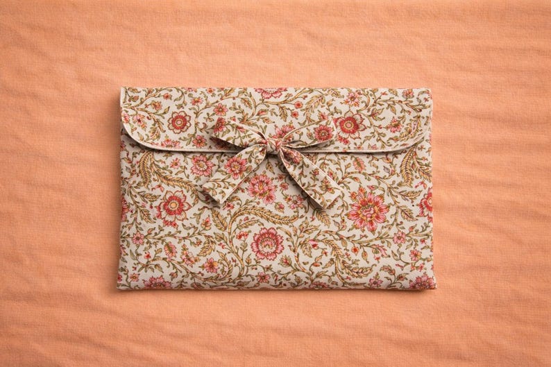 Op de afbeelding: Een clutch met bloemenpatroon en striksluiting. De clutch heeft een cr&egrave;mekleurige achtergrond met rode en roze bloemen en groene bladeren. De achtergrond is perzikkleurig.