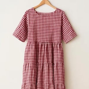 May include: A short-sleeved dress featuring a red and white gingham check pattern. The garment has a round neckline and a tiered skirt design with horizontal seams that create a gathered, flowing silhouette, hanging from a wooden hanger.