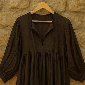 May include: A dark brown dress with a V-neck and long sleeves, suspended on a wooden hanger. The dress features a gathered waist and a flowing skirt, offering a relaxed fit. The fabric seems to be a soft, natural material.