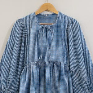 Może przedstawiać: Niebiesko-biała sukienka w kratę z długimi, pękatymi rękawami i wiązaniem przy szyi. Sukienka wisi na drewnianym wieszaku. Sukienka ma marszczoną talię i luźny krój.