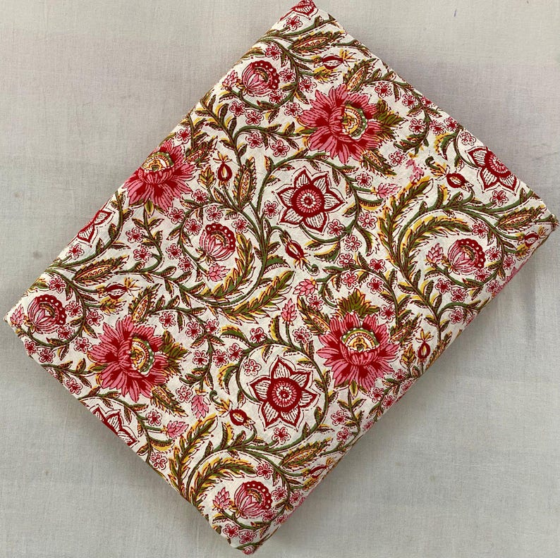 Op de afbeelding: Gevouwen stof met een bloemenpatroon. De stof heeft grote, roze en rode bloemen met groene bladeren en stelen op een witte achtergrond. Het bloemmotief wordt herhaald over de stof.