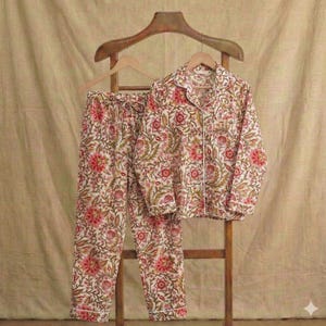 Op de afbeelding: Een tweedelige pyjamaset met een bloemenpatroon in rood, roze en groen op een cr&egrave;mekleurige achtergrond. De top met lange mouwen en de broek worden gepresenteerd op een houten hanger en standaard. De pyjama's hebben witte biezen.