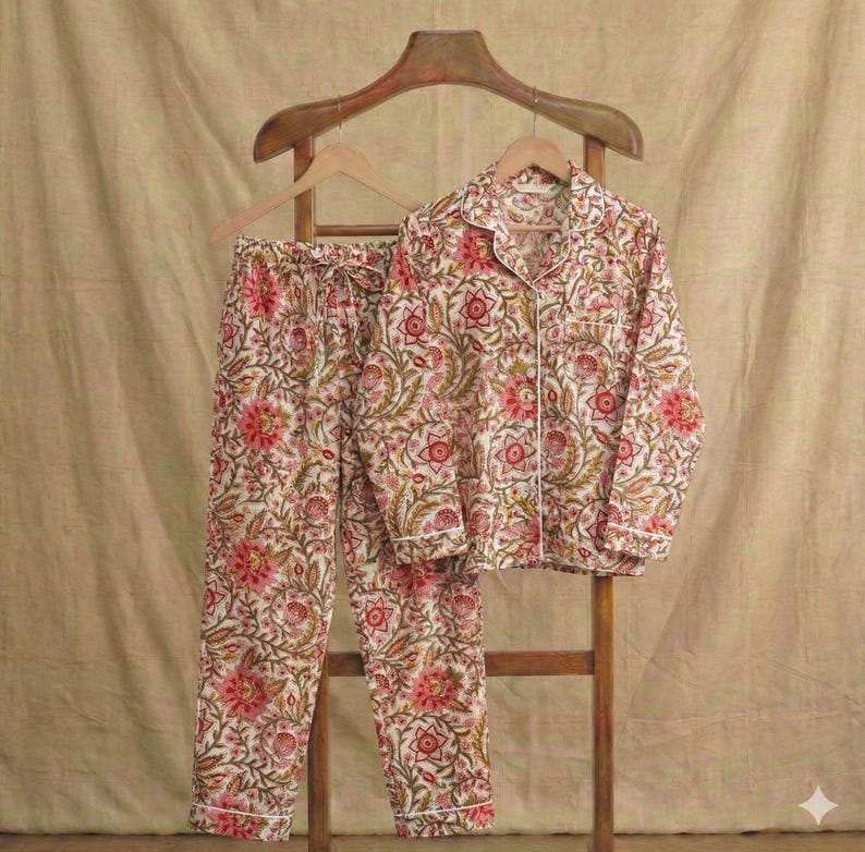 Op de afbeelding: Een tweedelige pyjamaset met een bloemenpatroon in rood, roze en groen op een cr&egrave;mekleurige achtergrond. De top met lange mouwen en de broek worden gepresenteerd op een houten hanger en standaard. De pyjama's hebben witte biezen.