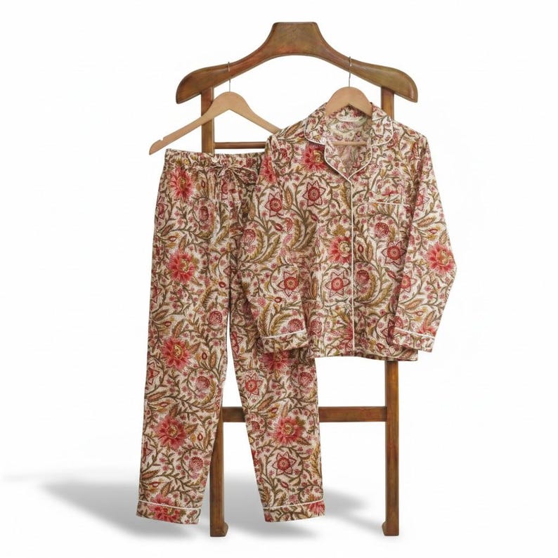 Op de afbeelding: Een tweedelige pyjamaset met een bloemenpatroon in rood, groen en cr&egrave;me. De top met lange mouwen en de broek worden gepresenteerd op een houten hanger en standaard. De pyjama heeft witte biezen.