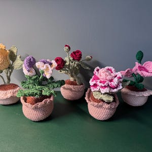 Pode incluir: Cinco arranjos de flores de crochê feitos à mão em vasos pequenos. As flores são de várias cores, incluindo amarelo, roxo, vermelho e rosa. Os vasos são de cor rosa claro com uma borda ondulada.