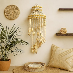 Puede incluir: Un colgante de pared decorativo con un diseño de conchas crema y amarillo. El colgante presenta un diseño en capas con detalles colgantes de conchas. También se ven una decoración de pared redonda tejida y una planta en maceta.