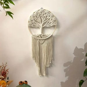 May include: A macrame wall hanging featuring a tree design. The tree is crafted with off-white rope, with a circular frame and long fringe. The tree's branches and roots are intricately knotted, creating a textured, natural aesthetic.