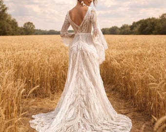 May include: A white, long-sleeved wedding dress with a low back and a flowing train. The dress features intricate lace detailing and bell sleeves. The bride is standing in a field of wheat.
