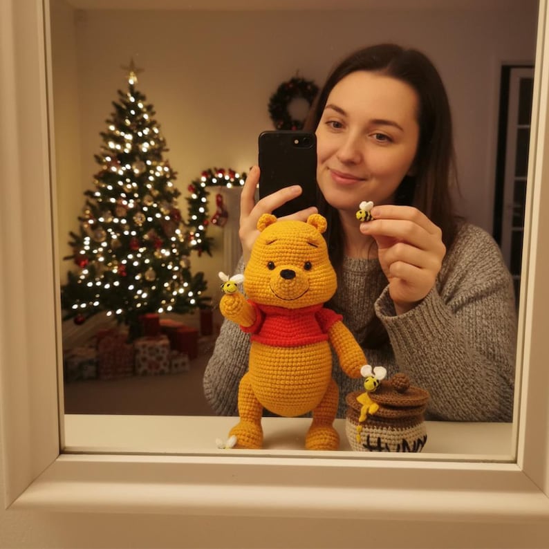 May include: A crocheted Winnie the Pooh figure with a red shirt and a honey pot. The figure is yellow with black eyes and a smiling face. A small crocheted bee is attached to the figure's hand. The honey pot is brown and white.