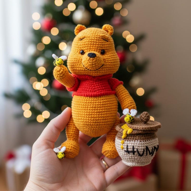 May include: A handmade crochet Winnie the Pooh plush toy. The yellow bear wears a red shirt and holds a bee. A small honey pot with the word "HUNNY" is next to the bear. The background is a Christmas tree.
