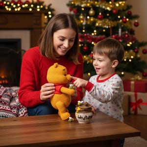 May include: A crocheted Winnie the Pooh doll in yellow with a red shirt, held by a person. A child points at the doll's nose. A crocheted honey pot with bees and the word "HUNNY" is on the table. Christmas tree in the background.