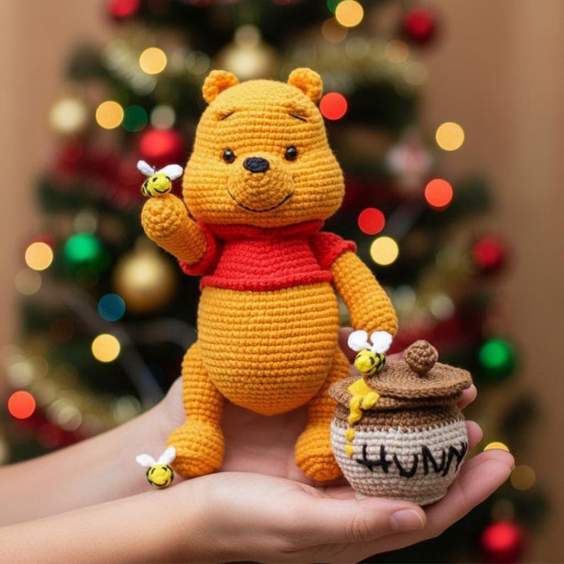 May include: A crocheted Winnie the Pooh doll in yellow and red, holding a bee and a honey pot. The honey pot is beige with the word "HUNNY" on it. The background is a blurred Christmas tree.