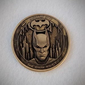 Può includere: Una moneta color oro con il cappuccio e il volto di Batman, con le scritte "Gotham City" e "The Dark Knight". La moneta presenta un design dettagliato con uno sfondo di paesaggio urbano e il simbolo di Batman.