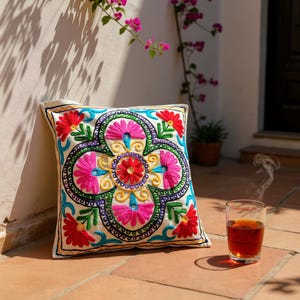 Peut inclure: Coussin carré décoratif avec broderie florale colorée. Le motif floral comprend des fils rouges, roses, jaunes, verts et bleus. Un verre de thé est posé à côté du coussin.