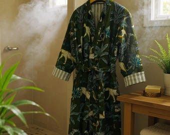 May include: A green robe with a jungle print featuring leopards and foliage hangs in a bathroom. The robe has a matching belt and striped cuffs. The bathroom has a wooden side table with towels and a plant.