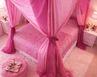 May include: A pink bedroom with a canopy bed draped in varying shades of pink fabric. The bed is covered with a patterned pink comforter. A pink ottoman with a tray of items sits on a fluffy white rug.