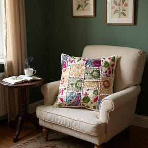 May include: A patchwork cushion with floral and paisley patterns in pink, blue, green, and gold. The cushion sits on a cream armchair. The room has dark green walls and a small wooden side table with a cup of tea.