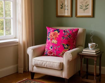 May include: A bright pink floral cushion rests on a cream-colored armchair. The cushion features a vibrant pattern of red, yellow, and blue flowers. Two framed botanical prints hang on the green wall, and a small table holds a teacup.