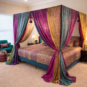 Op de afbeelding: Een hemelbed met levendige, bedrukte beddengoed en gedrapeerde gordijnen in tinten paars, goud en blauwgroen. Het bedframe is blauw en de kamer heeft een gezellige, uitnodigende sfeer met zachte verlichting.