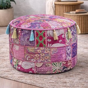 Puede incluir: Un puf redondo de patchwork en tonos rosa, morado y magenta. La parte superior y los lados están adornados con una variedad de cuadrados de tela estampada. Los bordes están ribeteados con un borde rosa y acentuados con borlas azules y amarillas. Este otomán decorativo mide unos 45 cm de diámetro.