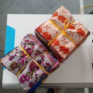 Może przedstawiać: Dwa stosy kwadratów materiału, każdy związany żółtą wstążką. Jeden stos ma kwiatowy wzór z różem, brązem i zielenią na jasnym tle. Drugi stos ma pomarańczowo-biały geometryczny wzór.