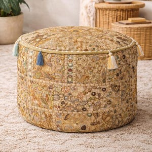 May include: Circular beige ottoman with a patchwork design showcasing floral and geometric patterns. The ottoman has decorative trim and is embellished with blue and yellow tassels. It rests on a beige rug, creating a bohemian aesthetic.