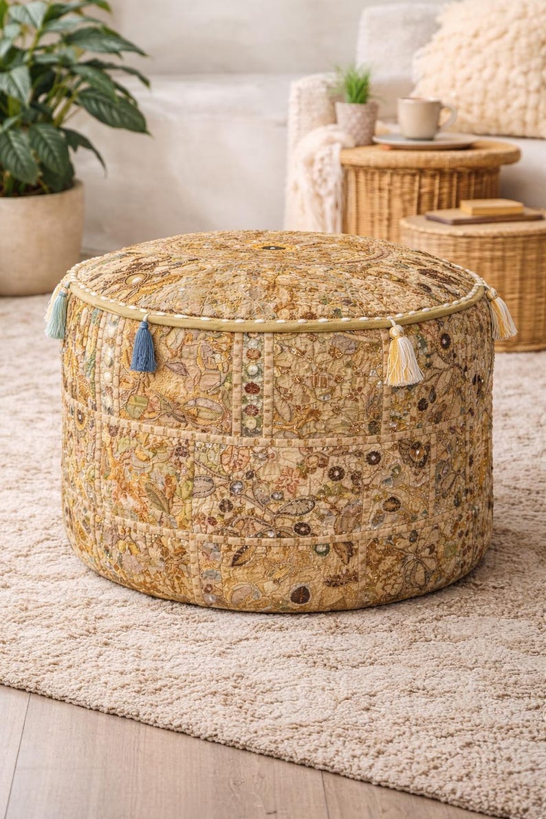 May include: Circular beige ottoman with a patchwork design showcasing floral and geometric patterns. The ottoman has decorative trim and is embellished with blue and yellow tassels. It rests on a beige rug, creating a bohemian aesthetic.