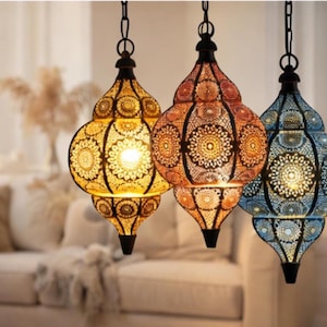 Puede incluir: Tres lámparas colgantes decorativas con intrincados diseños de metal calado. Las lámparas son de color amarillo, naranja y azul, cada una con una parte superior e inferior negra. Cuelgan de cadenas negras.