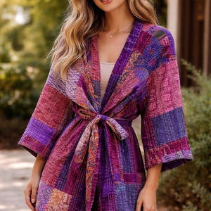 May include: A patchwork kimono-style robe in shades of purple and pink. The robe features a tie closure at the waist and three-quarter length sleeves. The fabric appears to be quilted with visible stitching.