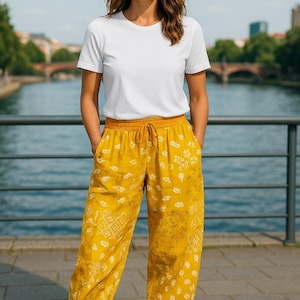 Op de afbeelding: Een persoon draagt een wit t-shirt en een gele broek met patroon en een taille met trekkoord. Ze draagt witte sneakers. De broek heeft een bloemenpatroon. De achtergrond omvat een rivier en een brug.