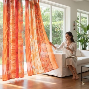 Puede incluir: Cortinas estampadas en naranja y rojo cuelgan frente a una ventana grande. Las cortinas tienen un diseño de patchwork con patrones florales y geométricos. La tela transparente permite el paso de la luz. Una mujer está sentada en un sofá blanco.
