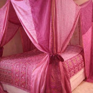 May include: A pink canopy bed with patterned bedding and draped fabric. The bedspread and canopy feature a repeating design. The canopy is tied, creating a romantic atmosphere. The room has a soft, warm colour palette.