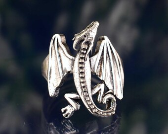 May include: A silver-colored dragon ring with detailed wings and a textured body. The dragon is depicted with its head raised, and its wings are spread. The ring is likely made of metal and has a polished finish. The ring is a fantasy-themed jewelry item.