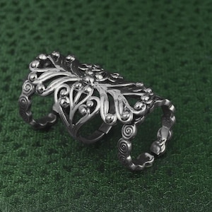 Könnte beinhalten: Silberfarbener, beweglicher Fingerring mit einem verzierten, floralen Design. Der Ring hat drei Bänder, die durch ein dekoratives Mittelstück mit aufwendigen Details verbunden sind. Das Design umfasst Wirbelmuster und kleine, erhabene Verzierungen, die eine Vintage-Ästhetik erzeugen.