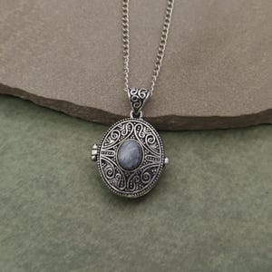 Peut inclure: Collier pendentif avec un médaillon ovale argenté. Le médaillon présente un motif orné et tourbillonnant autour d'une pierre centrale ovale bleu clair. Le collier a une chaîne délicate.