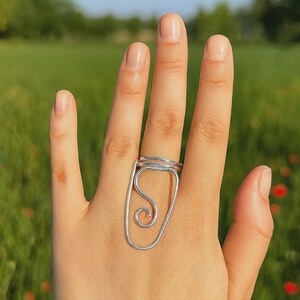 May include: A silver-colored wire ring with a unique design featuring a rectangular frame and an interior swirl. The ring is worn on a finger, showcasing its artistic and modern aesthetic. The background is a blurred green field and blue sky.