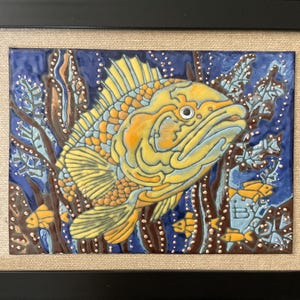 Può includere: Arte su piastrella di ceramica incorniciata raffigurante un pesce giallo e arancione con dettagli blu, su sfondo blu scuro con piante acquatiche. L'opera è incorniciata in una cornice di legno nera con un passe-partout neutro.