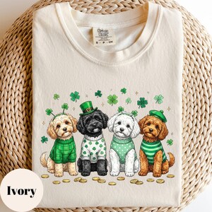 May include: Ivory-colored t-shirt featuring four cartoon dogs dressed in St. Patrick's Day attire. Each dog wears a green sweater or vest, and some have hats or headbands with shamrocks. Gold coins and shamrocks are scattered around the dogs.