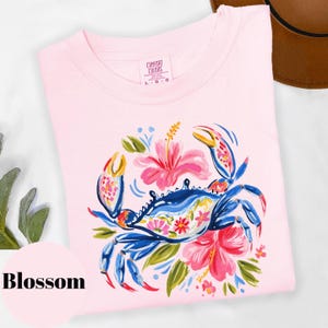Może przedstawiać: Jasnor&oacute;żowa bluza Comfort Colors z niebieskim krabem i kwiatowymi akcentami. Krab otoczony jest r&oacute;żowymi kwiatami hibiskusa i zielonymi liśćmi. Słowo "Blossom" znajduje się w r&oacute;żowym okręgu w lewym dolnym rogu.
