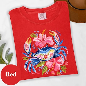 Może przedstawiać: Czerwona koszulka z ilustracją niebieskiego kraba i kwiatowymi detalami. Krab otoczony jest r&oacute;żowymi kwiatami hibiskusa i zielonymi liśćmi. Koszulka ma okrągły dekolt, a tekst "Red" znajduje się w czerwonym okręgu.