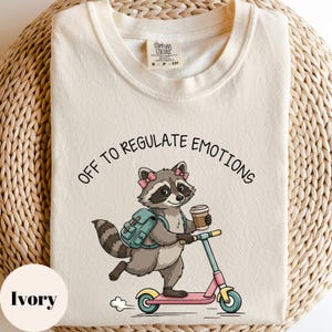 May include: Ivory-colored t-shirt with a raccoon graphic riding a pink scooter, holding a coffee cup, and wearing a backpack. The text "OFF TO REGULATE EMOTIONS" arches above the raccoon. The shirt has a crew neck.