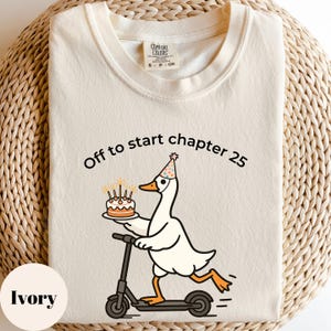 May include: Ivory-colored t-shirt with the text "Off to start chapter 25." A cartoon goose wearing a party hat and holding a cake rides a scooter. The shirt has a Comfort Colors tag.
