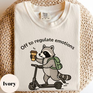 May include: Ivory-colored t-shirt with the text "Off to regulate emotions" above a cartoon raccoon riding a scooter and holding a coffee cup. The raccoon wears a green backpack. The shirt has a Comfort Colors tag.