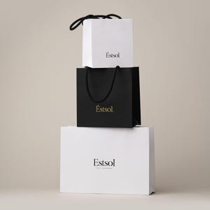 Peut inclure: Trois sacs &agrave; provisions empil&eacute;s. Le sac sup&eacute;rieur est blanc avec une poign&eacute;e noire et le mot "&Ecirc;stsol" imprim&eacute; en noir. Le sac du milieu est noir avec une poign&eacute;e noire et le mot "&Ecirc;stsol" imprim&eacute; en or. Le sac inf&eacute;rieur est blanc avec le mot "Estsol" imprim&eacute; en noir.