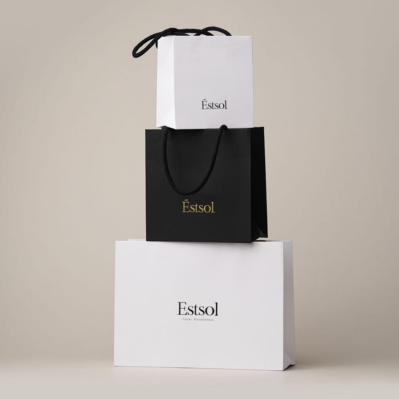Peut inclure: Trois sacs &agrave; provisions empil&eacute;s. Le sac sup&eacute;rieur est blanc avec une poign&eacute;e noire et le mot "&Ecirc;stsol" imprim&eacute; en noir. Le sac du milieu est noir avec une poign&eacute;e noire et le mot "&Ecirc;stsol" imprim&eacute; en or. Le sac inf&eacute;rieur est blanc avec le mot "Estsol" imprim&eacute; en noir.