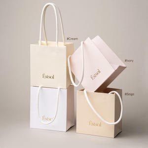 Peut inclure: Quatre petits sacs &agrave; provisions cr&egrave;me, ivoire, beige et blanc, avec des poign&eacute;es en corde blanche. Les sacs portent le mot "Estsol" imprim&eacute; en or. Le fond est de couleur neutre.