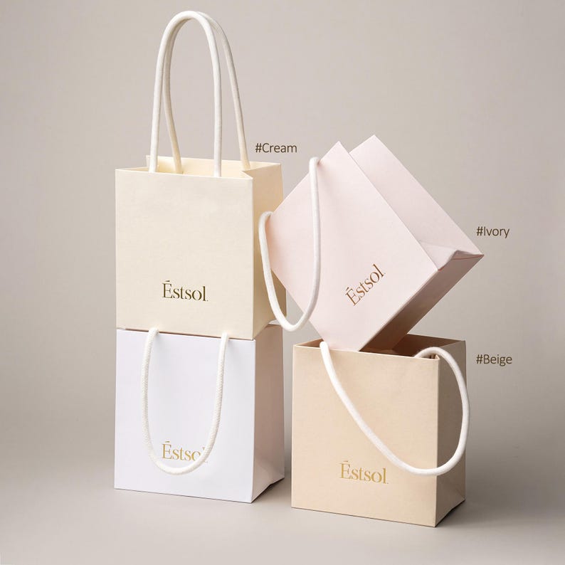 Peut inclure: Quatre petits sacs &agrave; provisions cr&egrave;me, ivoire, beige et blanc, avec des poign&eacute;es en corde blanche. Les sacs portent le mot "Estsol" imprim&eacute; en or. Le fond est de couleur neutre.