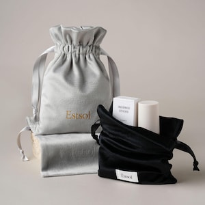 Pode incluir: Uma bolsa de veludo cinza-claro com cordão e o texto dourado Estsol, um pano retangular de veludo cinza a condizer e uma bolsa de veludo preta com cordão e uma etiqueta branca com o texto Estsol, contendo uma caixa e um tubo brancos.